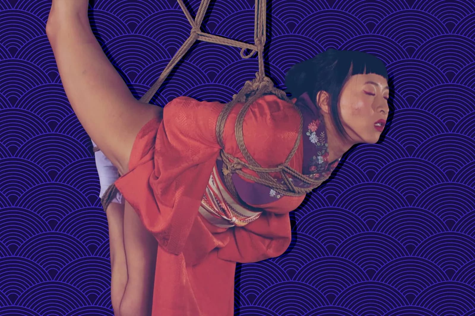 woman in a red kimono suspended in shibari rope, with a purple wave-patterned background.