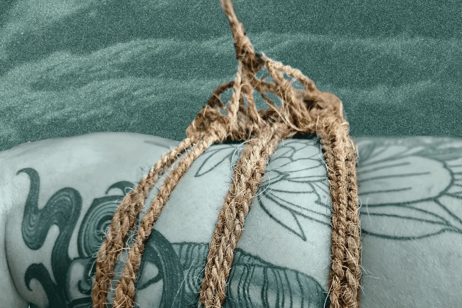 Tattooed leg bound in coconut rope with a leg harness, set against a textured teal background.