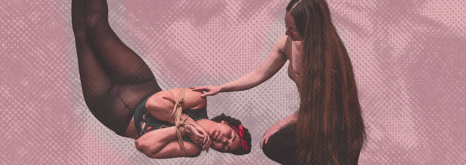 Person suspended in rope harness while another gently supports them against a pink backdrop