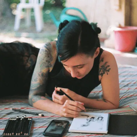 A person concentrating while drawing with visible arm tattoo and art supplies nearby.