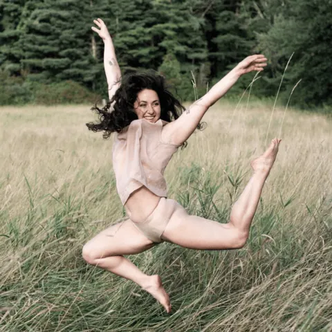 A person in mid-air leaping above a grassy field with evergreen trees.