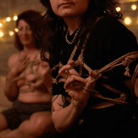 A person with visible tattoos tying shibari rope in a studio with warm lighting.
