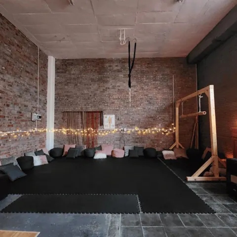 A rope studio interior with brick walls, suspended equipment, cushions, and warm lighting.